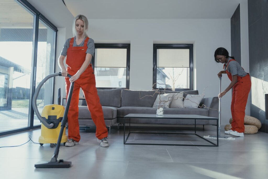 pexels photo 6195277 Two women in red overalls vacuuming and sweeping a modern living room.